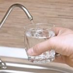 Hand holding a glass with clean water under faucet in modern kitchen; Zing Plumbing promotes water quality safety.