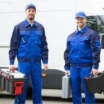 Two male Zing Plumbing workers in blue uniforms holding toolboxes beside a plumbing van.