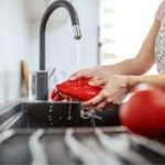Person washing a red bell pepper in a stainless steel sink, promoting cleanliness for effective plumbing maintenance.