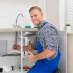 Smiling male plumber working under a clean kitchen sink, fixing plumbing pipes for Zing Plumbing service in Tucson.