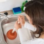 A woman unclogging a sink with a plunger while cleaning supplies are visible, promoting Zing Plumbing's maintenance tips.