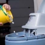 Person pouring items into a gray waste container, with a bright yellow bucket, in a clean disposal setting.