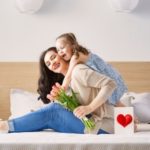 Smiling mother holding tulips and a heart card with child in a cozy bedroom. Zing Plumbing gift ideas.