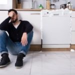 A man in casual attire sitting on a kitchen floor near a dishwasher with spilled liquid, representing plumbing issues.