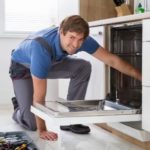 Male technician in a bright kitchen repairing a dishwasher, showcasing Zing Plumbing services. Toolbox on the floor.