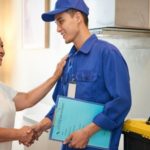 Delivery person in blue uniform gives folder to older woman in kitchen, highlighting professionalism in plumbing services.