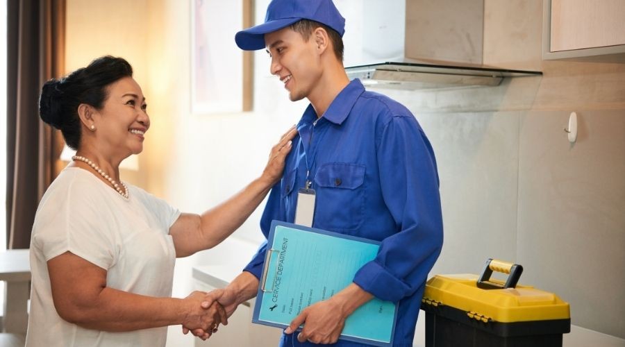 Delivery person in blue uniform shakes hands with older woman, discussing plumbing documents in modern indoor setting.