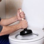 A person using a black plunger on a white toilet in a tiled bathroom, illustrating clogged toilet repair by Zing Plumbing.