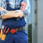 Repairman in blue overalls and plaid shirt, confidently posing outdoors, representing Zing Plumbing services.