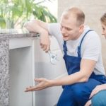 Male technician explains plumbing services to female client at kitchen counter. Zing Plumbing.