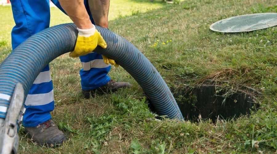 Worker in blue attire using a hose for plumbing maintenance in a grassy area. Zing Plumbing services in action.