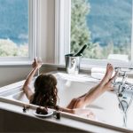 Person relaxing in a bathtub with champagne and towels, showcasing a spa-like bathroom experience by Zing Plumbing.