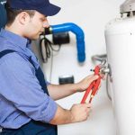 Male plumber in work attire using an adjustable wrench near a water heater at Zing Plumbing’s organized workspace.