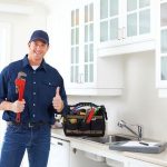 Smiling male plumber in blue uniform with adjustable wrench by kitchen sink, ready for plumbing services in Tucson.