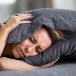 A woman in distress holding a gray pillow in a home setting, representing anxiety about plumbing issues like water hammer.