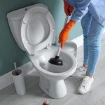 Person in orange gloves plunging a toilet in a modern bathroom, promoting Zing Plumbing's holiday plumbing maintenance tips.