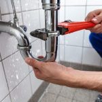 A person using a red wrench to fix a leaking pipe under a sink, showcasing Zing Plumbing's drain cleaning services.