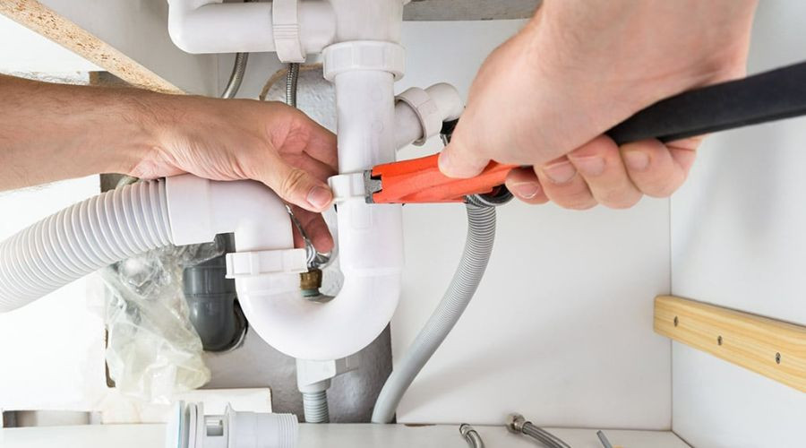 Hands working on plumbing connections under a sink with a pipe wrench, PVC pipes, and fittings at Zing Plumbing service.