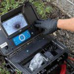 Portable inspection camera for plumbing maintenance, showing underground structures on screen, operated by a gloved hand.